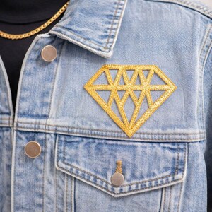 Puede incluir: Chaqueta vaquera azul claro con un parche dorado en forma de diamante en el pecho. La chaqueta tiene un diseño clásico con botones y bolsillos. Se ve un collar de cadena dorada.