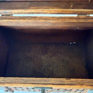 May include: Open wooden chest with a woven front panel. The chest is made of dark brown wood with visible grain and a hinged lid. The interior is empty, revealing the natural wood color. The front panel has a woven design with metal hardware.