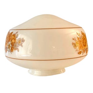 May include: Cream-colored glass lampshade with floral designs in shades of orange and yellow. The lampshade has a rounded shape with a brown trim and a small base. The floral pattern is repeated on both sides of the lampshade.