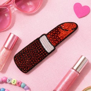 Può includere: Una toppa a forma di rossetto rosso con contorno nero e dettagli bianchi. Il rossetto è impreziosito da paillettes rosse. L'immagine include anche occhiali da sole rosa, lucidalabbra e un ritaglio a forma di cuore, il tutto su sfondo rosa.