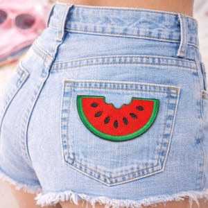 以下が含まれることがあります： バックポケットに赤と緑のスイカのパッチが付いた、明るい青色のデニムショーツ。 スイカのパッチは一口食べられており、黒い種が見えています。 ショーツはほつれた裾になっています。