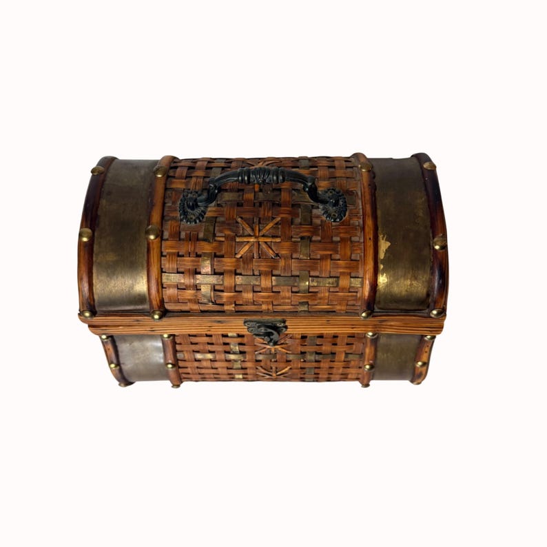 May include: A small, decorative chest with a woven bamboo body, brass-colored metal accents, and a black handle. The chest features a starburst design on the top and is secured with a latch. The overall design is reminiscent of a treasure chest.