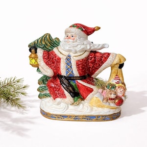 May include: A ceramic Santa Claus figurine, dressed in a red and white suit with gold accents, carrying a green Christmas tree and a sack of toys. The figurine has a white beard and a red hat. A yellow lantern is in his left hand.