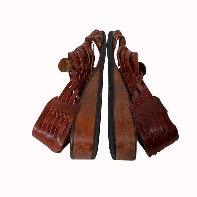 May include: Pair of brown leather sandals with woven straps and wooden wedge heels. The sandals have a button closure and a black sole. The shoes are displayed against a white background.