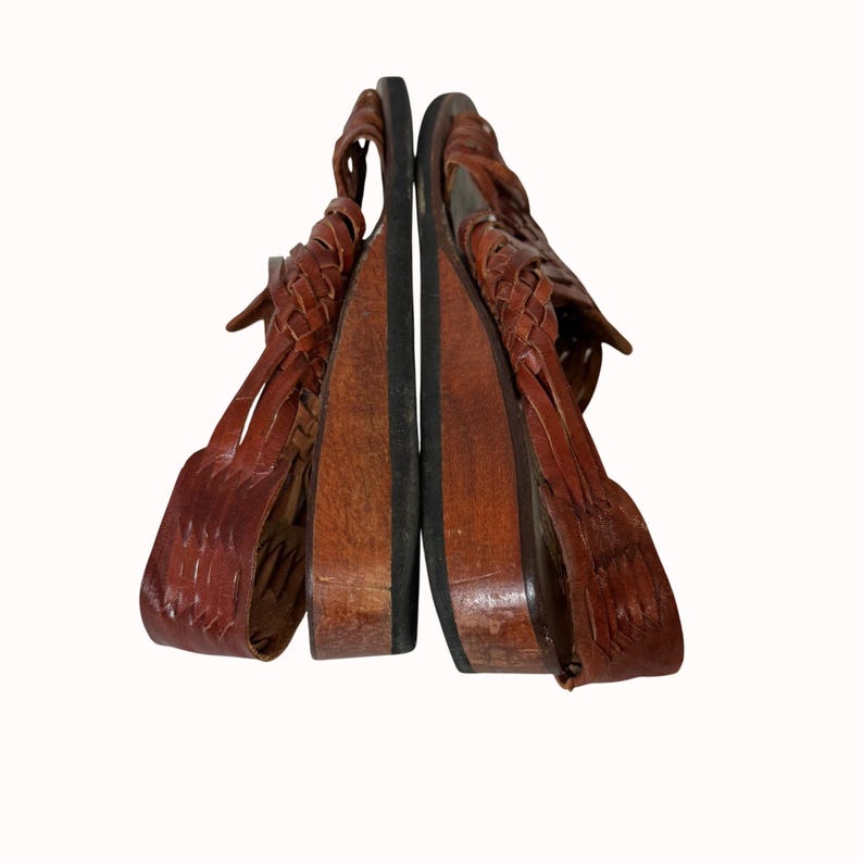 May include: A pair of brown leather sandals with woven straps and wooden soles. The sandals are displayed side by side, showcasing the intricate weaving detail and the natural wood color of the soles.