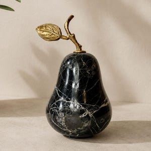 May include: A pear-shaped decorative object crafted from polished black marble with prominent white veining. The top of the pear features a textured, metallic gold-toned stem and a single leaf extending upwards from the center of the fruit.