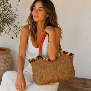 Puede incluir: Un bolso tote tejido marrón con asas naranjas y un ribete con estampado floral colorido. El bolso es de color marrón natural y parece estar hecho de paja o un material similar. El bolso lo sostiene una persona.