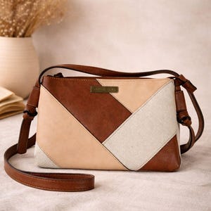 Puede incluir: Un bolso bandolera marrón, beige y crema con un diseño geométrico. El bolso tiene una correa larga y ajustable y una pequeña etiqueta rectangular con texto. El bolso está hecho de cuero y lona.