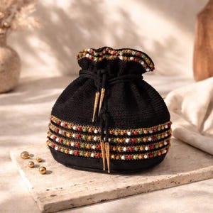 May include: Black drawstring pouch with beaded embellishments. The pouch features rows of colorful beads in red, green, white, and gold. Gold-colored metal accents and a drawstring closure complete the design. The pouch is sitting on a stone surface.