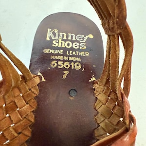 May include: Close-up of a brown leather sandal with woven straps. The insole is stamped with "Kinney Shoes, Genuine Leather, Made in India" and the size 7. The sandal has a vintage aesthetic.