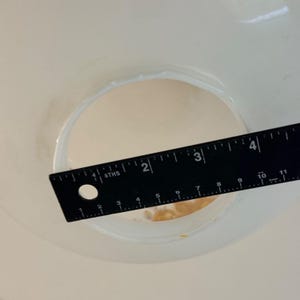 May include: A black ruler with white markings is placed over a white circular opening. The ruler shows measurements in inches, with the opening measuring approximately 2 inches in diameter. The image is taken from a top-down perspective.
