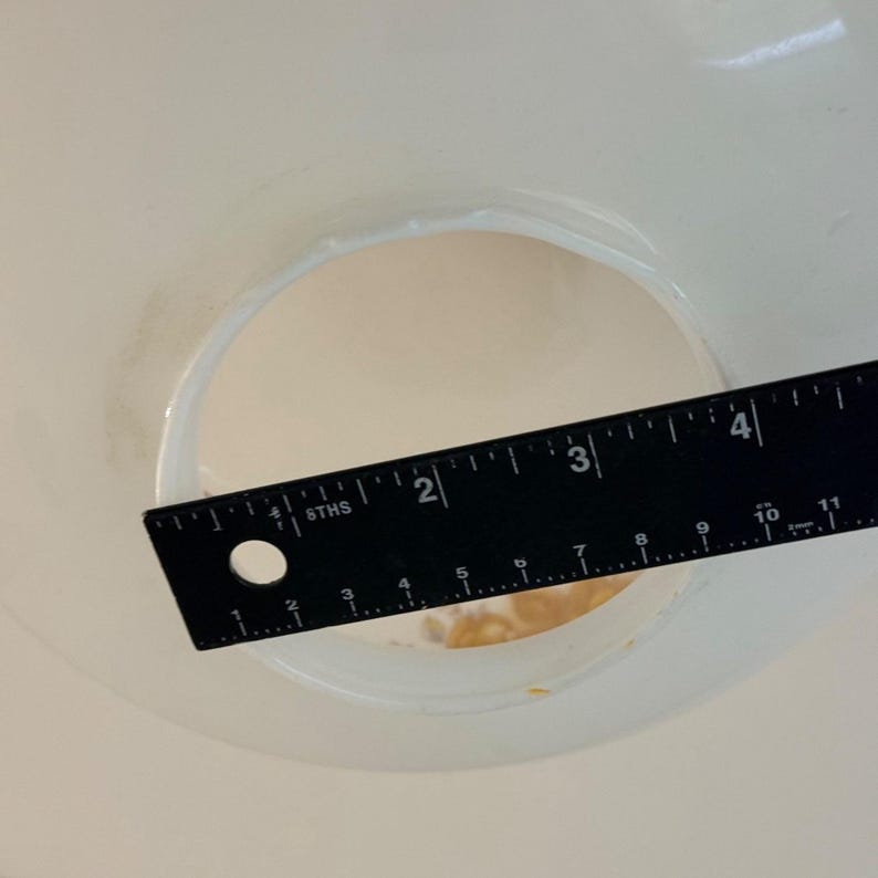May include: A black ruler with white markings is placed over a white circular opening. The ruler shows measurements in inches, with the opening measuring approximately 2 inches in diameter. The image is taken from a top-down perspective.