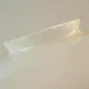 May include: A close-up of a light-colored, possibly ceramic, object with a curved, wing-like shape. The object has a smooth, glossy surface with subtle variations in tone, suggesting a handmade or natural aesthetic. The lighting creates highlights and shadows.