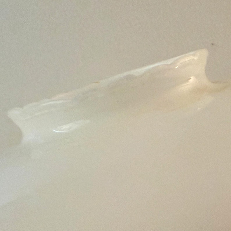 May include: A close-up of a light-colored, possibly ceramic, object with a curved, wing-like shape. The object has a smooth, glossy surface with subtle variations in tone, suggesting a handmade or natural aesthetic. The lighting creates highlights and shadows.