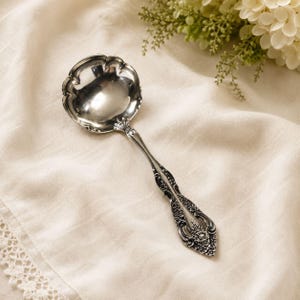 May include: A silver-toned metal ladle featuring a round, scalloped bowl and a handle decorated with intricate, embossed floral and scrollwork patterns. The polished surface of the bowl reflects light, while the handle displays dark, recessed details.