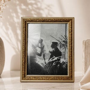 May include: A rectangular picture frame with an ornate, embossed gold-toned border. The frame contains a black and white photograph depicting two figures near a window with bright light streaming in, alongside a potted plant with large leaves.