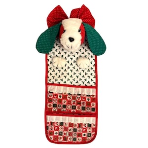 以下が含まれることがあります： 犬の形をしたクリスマスをテーマにした吊り下げ式オーガナイザー。犬は白いぬいぐるみのような顔、赤と緑の耳、赤いリボンをしています。オーガナイザーには、赤、緑、白のフェスティバルパターンで、レースでトリミングされたポケットが付いています。