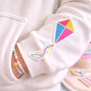 Puede incluir: Una sudadera con capucha blanca con un parche de cometa colorido en la manga. El diseño de la cometa presenta secciones rojas, azules, amarillas y rosas con un borde blanco. Una pulsera con cuentas pastel y un dije de carita sonriente es visible en el bolsillo.