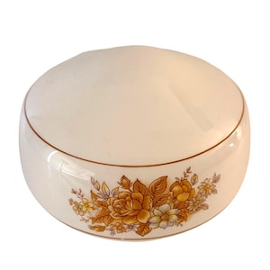 May include: Cream-colored ceramic container with a rounded lid and a floral design. The container features a band of brown trim and a detailed pattern of yellow, orange, and white flowers and leaves around the base.