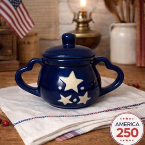 May include: A navy blue ceramic sugar bowl with a lid and heart-shaped handles. The bowl features three cream-coloured stars and sits on a white cloth. The image includes an American flag and an "America 250" emblem.
