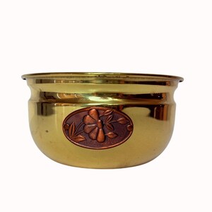 May include: A brass-coloured planter with a copper-toned floral design. The planter has a rounded shape with a slightly flared rim. The floral design is oval-shaped and features a flower and leaves.