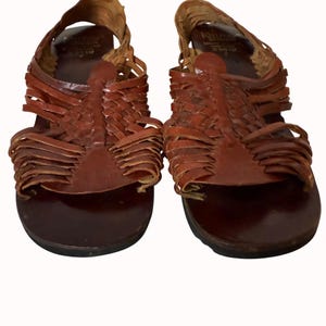 May include: A pair of brown leather sandals with woven straps. The sandals have a dark brown sole and a woven design across the top of the foot. The straps are made of thin strips of leather. The sandals are displayed on a white background.