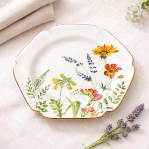 May include: A white ceramic plate with a scalloped edge and a gold-toned rim, featuring a colorful watercolor-style illustration of various wildflowers including yellow, orange, and purple blossoms with green stems and leaves.