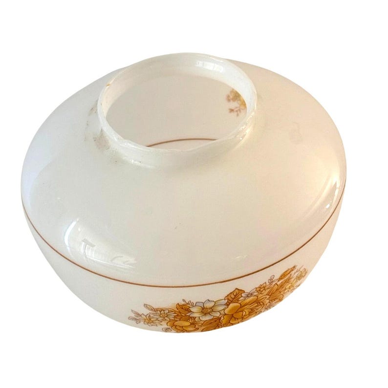 May include: A vintage, off-white glass lampshade with a rounded shape and a wide opening. The shade features a decorative band of yellow and brown floral patterns around the base. A thin brown line accents the design.