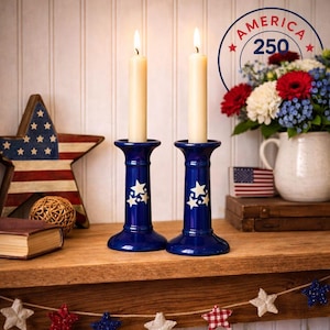 May include: Two lit candles in blue ceramic candle holders with star patterns. The holders are on a wooden shelf with patriotic decorations, including a star-shaped flag and a floral arrangement. The image includes the text "America 250".