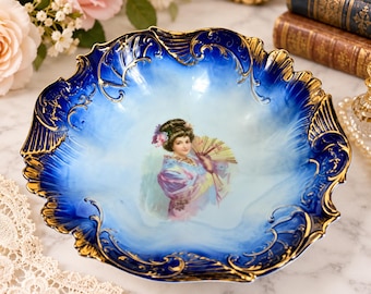 May include: A decorative porcelain bowl featuring a deep blue gradient rim with ornate gold filigree detailing. The center of the bowl displays a painted illustration of a person with dark hair holding a yellow fan, wearing a purple garment.