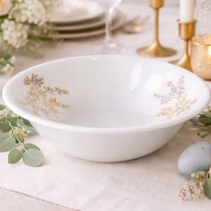 Op de afbeelding: Een witte keramische serveerschaal met een gouden bloemenontwerp. De kom heeft een brede, ondiepe vorm en is versierd met delicate goudkleurige botanische accenten. De kom staat op een wit tafelkleed, met ander serviesgoed op de achtergrond.