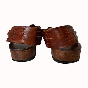 May include: A pair of brown leather platform sandals. The sandals have a wooden sole and a woven leather strap across the top of the foot. The sandals are viewed from the back.