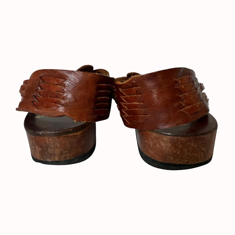 May include: A pair of brown leather platform sandals. The sandals have a wooden sole and a woven leather strap across the top of the foot. The sandals are viewed from the back.