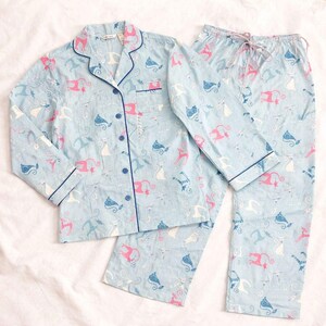 May include: Light blue pajama set with a cat pattern. The button-down top and pants feature a repeating design of pink, white, and blue cats. The set has navy blue trim and a chest pocket on the top. The pants have a drawstring waist.