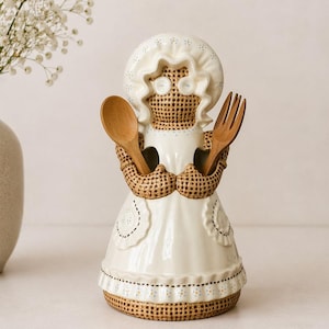 Peut inclure: Un porte-ustensiles de cuisine en céramique en forme de femme portant un bonnet et un tablier. Le support a une texture tissée sur les bras et la base, et tient une cuillère et une fourchette en bois. Le tablier est blanc avec des coutures décoratives.