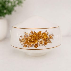 May include: A white ceramic bowl with a lid, featuring a floral design in shades of gold and white. The bowl has a rounded shape with a gold trim. The floral pattern includes roses and other flowers. A decorative piece for the home.
