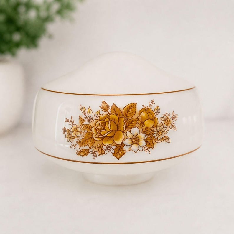 May include: A white ceramic bowl with a lid, featuring a floral design in shades of gold and white. The bowl has a rounded shape with a gold trim. The floral pattern includes roses and other flowers. A decorative piece for the home.