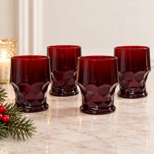 May include: A set of four deep red glass tumblers featuring a textured honeycomb pattern on the lower half. Each glass has a cylindrical shape with a smooth rim and a thick base, reflecting light through the translucent, dark crimson coloured glass.