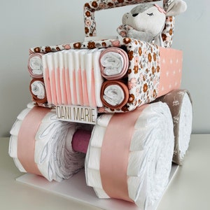 May include: A pink and brown diaper cake shaped like a jeep with a stuffed animal in the driver's seat. The cake is decorated with a floral pattern and the name "Dani Marie" is written on the front.