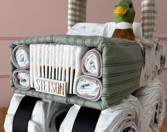 May include: A diaper cake shaped like a truck, featuring rolled diapers as wheels with black ribbon accents. The front grille is made of folded diapers with a white sign reading STETSON. A plush mallard duck sits in the driver area, which has a beige and white checkered roof.