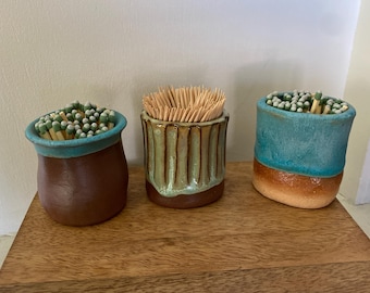May include: Three ceramic containers holding wooden items. The left is brown with a teal rim filled with matches. The center is a ribbed tan and green vessel holding toothpicks. The right is a teal and orange pot filled with matches.