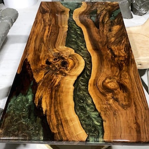 Könnte beinhalten: Eine Tischplatte aus Holz mit einem grünen Epoxidharzfluss, der durch die Mitte verläuft. Das Holz hat eine satte braune Farbe und ein natürliches Maserungsmuster.