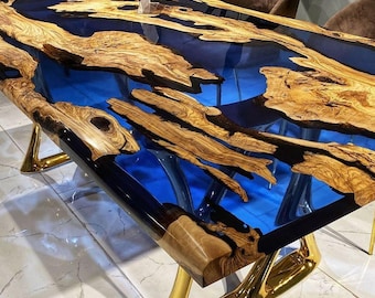 May include: A large, rectangular dining table with a unique epoxy resin top. The resin is a deep blue color and flows through the natural wood grain of the table top. The table is supported by four gold metal legs.