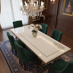 Könnte beinhalten: Ein formelles Esszimmer mit einem großen, rechteckigen weißen Tisch mit goldenen Verzierungen, umgeben von sechs grünen Samtstühlen. Ein Kristall-Kronleuchter hängt darüber, und ein gemusterter Teppich liegt unter dem Tisch. Kunstwerke und dunkle Holzverkleidungen vervollständigen die elegante Einrichtung.