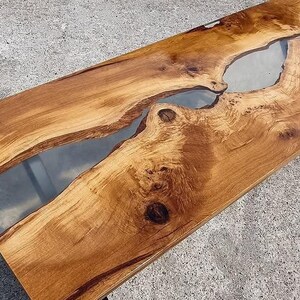 Könnte beinhalten: Ein langer Holztisch mit einer natürlichen Holzoptik und einem durchgehenden Fluss aus klarem Epoxidharz. Der Tisch hat ein schwarzes Metallgestell.