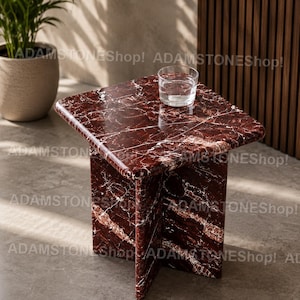 Könnte beinhalten: Ein quadratischer, roter Marmor-Beistelltisch mit weißen Adern. Der Tisch hat einen soliden Sockel und eine glatte, polierte Oberfläche. Ein klares Glas mit Wasser steht darauf. Der Tisch ist etwa 46 cm hoch.