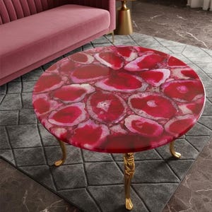 May include: A round coffee table with a vibrant red agate stone top. The table has a glossy finish and is supported by three ornate gold-colored legs. The table is set on a gray patterned rug.