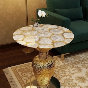 Könnte beinhalten: Ein runder Beistelltisch mit goldfarbenem, strukturiertem Sockel und einer Tischplatte aus weißen und goldfarbenen Steinen. Der Tisch steht auf einem gemusterten Teppich, im Hintergrund ein grünes Sofa. Eine kleine Vase mit einer weißen Blume steht auf dem Tisch.