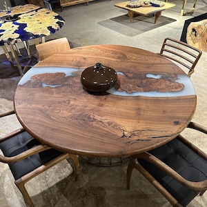 May include: A round wooden dining table with a light blue resin river design. The table is surrounded by four wooden chairs with black cushions. A dark brown decorative bowl sits on the table's surface. The table's diameter is approximately 127 cm.