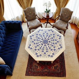 May include: An octagonal white coffee table with a blue floral design, set on a red rug. Two patterned armchairs and a small round table with a plant are visible. A blue sofa is on the left.